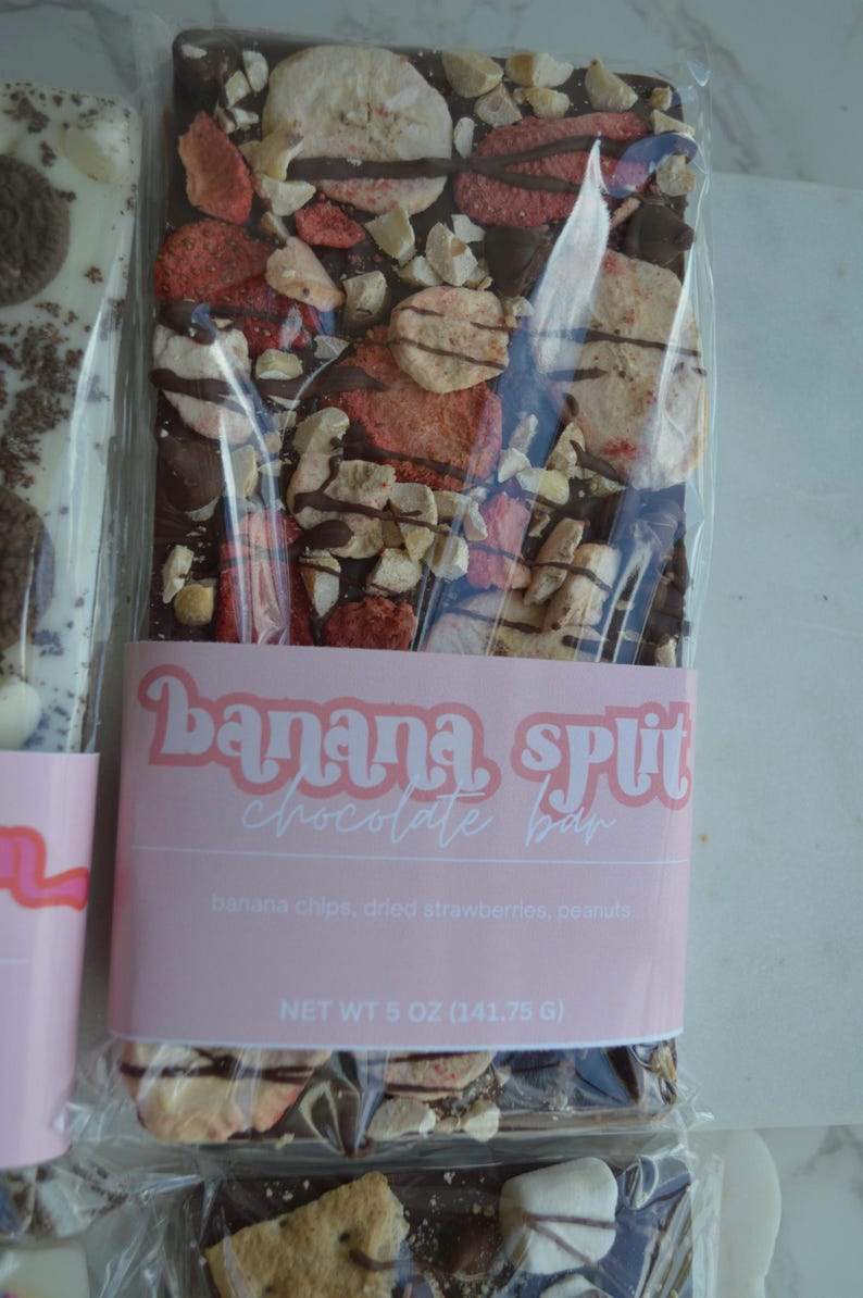 Peut inclure: Une barre de chocolat avec des chips de banane, des fraises s&eacute;ch&eacute;es et des cacahu&egrave;tes. La barre est emball&eacute;e dans du plastique transparent avec une &eacute;tiquette rose indiquant "Banana Split Chocolate Bar". Poids net 141,75 g.