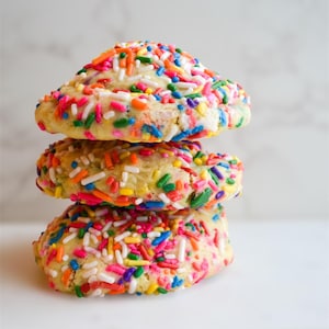 May include: A stack of three sprinkle cookies. The cookies are light yellow and covered in colorful sprinkles. The sprinkles are red, orange, yellow, green, blue, and white. The cookies are stacked on top of each other.