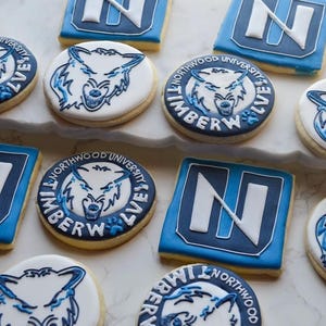 May include: A selection of iced sugar biscuits, decorated with blue and white icing. Some are round with a wolf logo and the words "Northwood University Timberwolves". Others are square with a white "N" on a blue background.