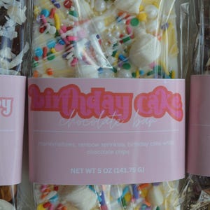 Peut inclure: Une barre de chocolat blanc avec des guimauves et des vermicelles multicolores, emball&eacute;e dans du plastique transparent. L'&eacute;tiquette rose indique "Birthday Cake Chocolate Bar". Poids net 141,75 g.