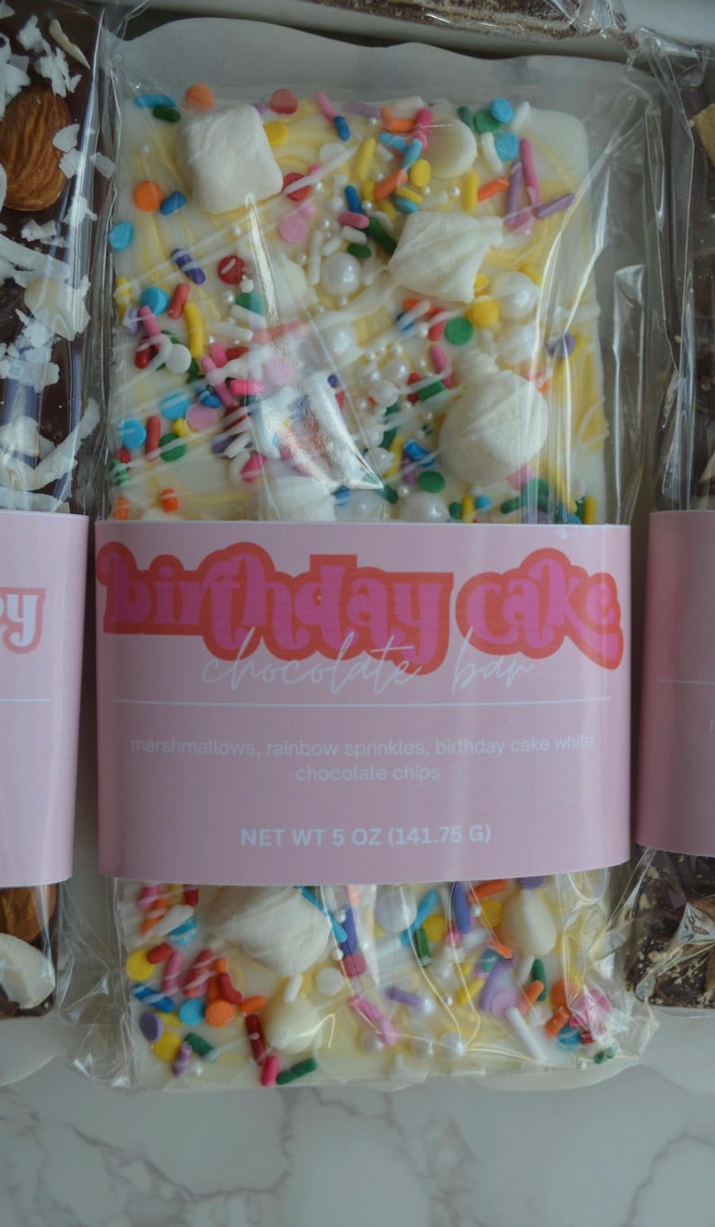 Peut inclure: Une barre de chocolat blanc avec des guimauves et des vermicelles multicolores, emball&eacute;e dans du plastique transparent. L'&eacute;tiquette rose indique "Birthday Cake Chocolate Bar". Poids net 141,75 g.