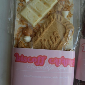 Peut inclure: Une barre de chocolat blanc avec des biscuits Biscoff, du caramel et un morceau de chocolat blanc. La barre est emball&eacute;e dans du plastique transparent avec une &eacute;tiquette rose indiquant "Biscoff Caramel Chocolate Bar". Le poids net est de 141,75 g.