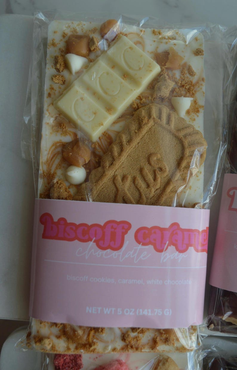 Peut inclure: Une barre de chocolat blanc avec des biscuits Biscoff, du caramel et un morceau de chocolat blanc. La barre est emball&eacute;e dans du plastique transparent avec une &eacute;tiquette rose indiquant "Biscoff Caramel Chocolate Bar". Le poids net est de 141,75 g.