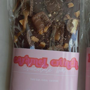 Peut inclure: Une barre de chocolat noir au caramel croquant dans un emballage transparent. Garnie de morceaux de Twix, Rolos et caramel. Une &eacute;tiquette rose indique "Caramel Crunch Chocolate Bar". Poids net : 141,75 g.