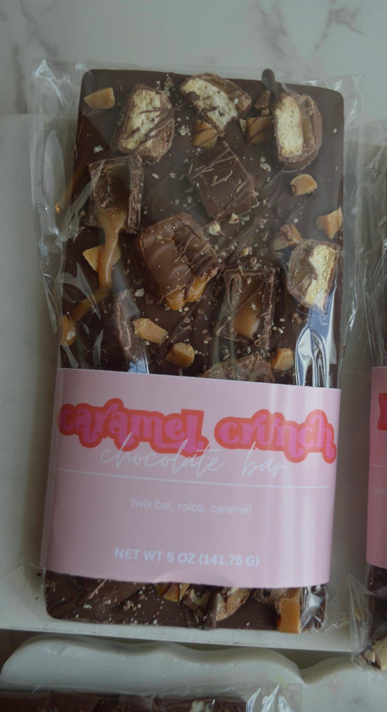 Peut inclure: Une barre de chocolat noir au caramel croquant dans un emballage transparent. Garnie de morceaux de Twix, Rolos et caramel. Une &eacute;tiquette rose indique "Caramel Crunch Chocolate Bar". Poids net : 141,75 g.