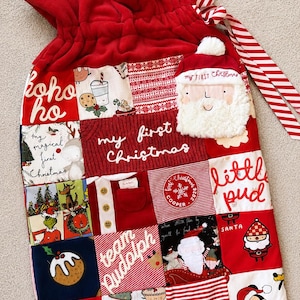 May include: A red drawstring gift bag featuring a patchwork design of various Christmas-themed fabrics. The bag includes text such as ho ho ho, my first Christmas, little pud, team Rudolph, and Santa, along with illustrations of Santa and Mickey Mouse.