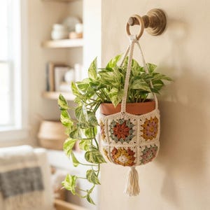 Puede incluir: Un soporte colgante para plantas hecho de cuadrados de ganchillo color crema en tonos apagados de óxido, verde salvia y amarillo mostaza. El soporte contiene una maceta de terracota con una planta pothos variegada y una borla.