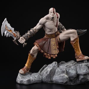 Peut inclure: Figurine détaillée d'un personnage musclé dans une pose dynamique, tenant deux armes à lames. La figurine est torse nu, avec une armure marron et dorée et une jupe marron. La figurine est posée sur un socle rocheux gris sur fond noir.