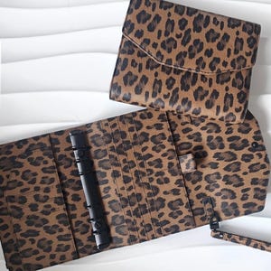 Könnte beinhalten: Ein braun-schwarzer Planer mit Leopardenmuster und eine passende Clutch werden offen und geschlossen gezeigt. Der Planer hat einen schwarzen Bindemechanismus und mehrere Kartenfächer. Die Clutch hat einen Klappenverschluss. Die Artikel befinden sich auf einer weißen Oberfläche.