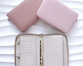 May include: Two closed rectangular zipped wallets in dusty rose and light pink, positioned above one open beige wallet featuring a gold-toned six-ring binder mechanism, multiple card slots, and a pen loop on the interior panels.