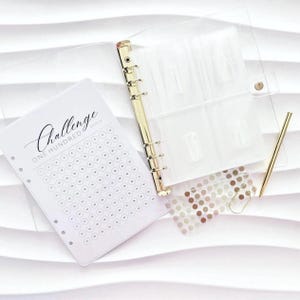 Könnte beinhalten: Ein transparenter Ordner mit goldenen Akzenten, ein goldener Stift und ein weißes Papier mit dem Aufdruck "Challenge". Das Papier hat ein Raster aus Kreisen zur Fortschrittsverfolgung. Eine transparente Folie mit braunen und goldenen Punkten und eine goldene Büroklammer sind ebenfalls enthalten.