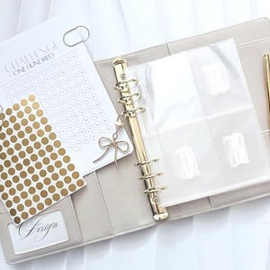 Könnte beinhalten: Ein beigefarbener Ringbuchplaner mit sechs Ringen und goldfarbenen Beschlägen, einem goldenen Stift und einer transparenten Einlage mit vier Fächern. Enthalten sind ein Blatt mit der Aufschrift CHALLENGE ONE HUNDRED, goldene Aufkleber und ein Anhänger.