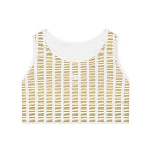 Op de afbeelding: Een wit crop top met een patroon van dunne, horizontale, mosterdgele stippellijnen gerangschikt in verticale kolommen. Het kledingstuk heeft een ronde halslijn en een klein, wit, rond geborduurd logo in het midden op de borst.