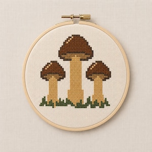 May include: A circular wooden embroidery hoop containing a cross-stitch pattern of three mushrooms. The mushrooms have dark brown caps and light tan stems, positioned above a row of dark green grass stitches on a cream-coloured fabric background.