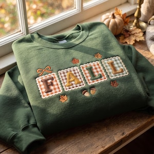 Puede incluir: Una sudadera de cuello redondo color verde bosque con la palabra FALL bordada en letras grandes. Cada letra está sobre un parche cuadrado con borde festoneado y estampado de cuadros vichy. El diseño incluye hojas naranjas y dos bellotas.
