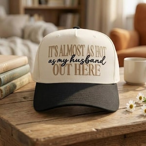 May include: A cream and black baseball cap with the embroidered text "IT'S ALMOST AS HOT as my husband OUT HERE". The cap is on a wooden table. The text is in brown and black lettering.