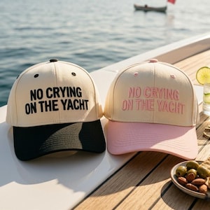 Puede incluir: Dos gorras de béisbol con el texto NO CRYING ON THE YACHT. La gorra izquierda es color crema con texto negro y visera negra. La gorra derecha es color crema con texto rosa y visera rosa. Ambas gorras tienen una corona estructurada.