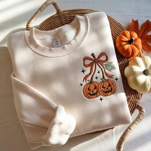 May include: A cream-colored crewneck sweatshirt featuring an embroidered design of two orange jack-o'-lanterns connected by a large orange bow with a green leaf. The design is accented with small black star shapes, creating a festive seasonal look.