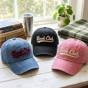May include: Three baseball caps in blue, black, and pink, each featuring the text Book Club embroidered on the front. The blue cap has red text, the black cap has white text, and the pink cap has white text. All caps have a distressed finish.