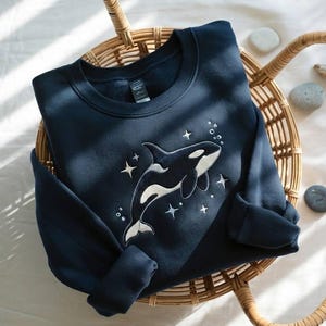 May include: A dark navy blue crewneck sweatshirt featuring an embroidered design of an orca whale surrounded by white stitched stars and small bubbles. The sweatshirt has long sleeves and a ribbed neckline, presented in a woven rattan basket.