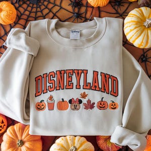 May include: A beige Disneyland sweatshirt with orange lettering and graphics. The word "DISNEYLAND" is arched across the chest. Below are pumpkin and fall-themed graphics, including a Minnie Mouse pumpkin and maple leaves. The sweatshirt is surrounded by pumpkins.