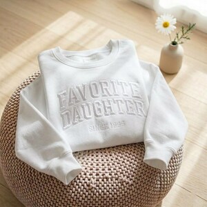 May include: A white sweatshirt with the embroidered text "FAVORITE DAUGHTER SINCE 1999". The crew neck sweatshirt is folded on a woven, round ottoman. A small vase with a daisy is in the background.