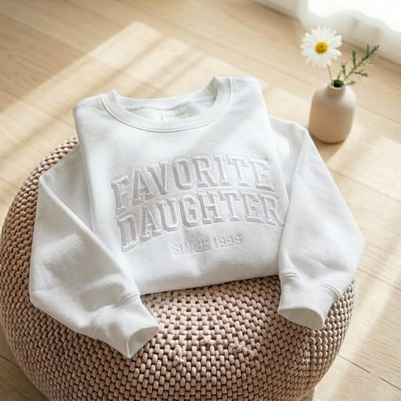May include: A white sweatshirt with the embroidered text "FAVORITE DAUGHTER SINCE 1999". The crew neck sweatshirt is folded on a woven, round ottoman. A small vase with a daisy is in the background.