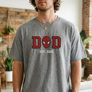 Pu&ograve; includere: Maglietta grigia con grafica rossa e nera con la scritta "DOD", una maschera di Spider-Man al centro e "EST. 2026" sotto. La maglietta &egrave; realizzata in un materiale morbido.