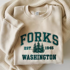May include: Cream-colored sweatshirt with the word "FORKS" in teal lettering. Below the word "FORKS" are three embroidered pine trees, the text "EST. 1945" and the word "WASHINGTON". The sweatshirt is folded on a cream-colored blanket.