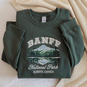 May include: A dark green crewneck sweatshirt featuring an embroidered graphic of a mountain range, pine trees, and a lake. The text reads BANFF National Park ALBERTA, CANADA in white and light blue thread with a white interior neck label.