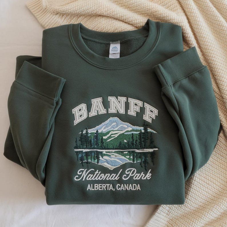 May include: A dark green crewneck sweatshirt featuring an embroidered graphic of a mountain range, pine trees, and a lake. The text reads BANFF National Park ALBERTA, CANADA in white and light blue thread with a white interior neck label.