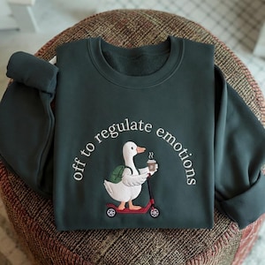May include: A dark green sweatshirt with the embroidered text "off to regulate emotions." The design shows a white duck on a red scooter, carrying a coffee cup and a backpack. The sweatshirt is displayed on a patterned surface.
