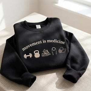 May include: Black sweatshirt with the phrase "movement is medicine" embroidered in white. Below the text are embroidered icons of a dumbbell, kettlebell, sneakers, jump rope, and yoga mat. The sweatshirt is folded.