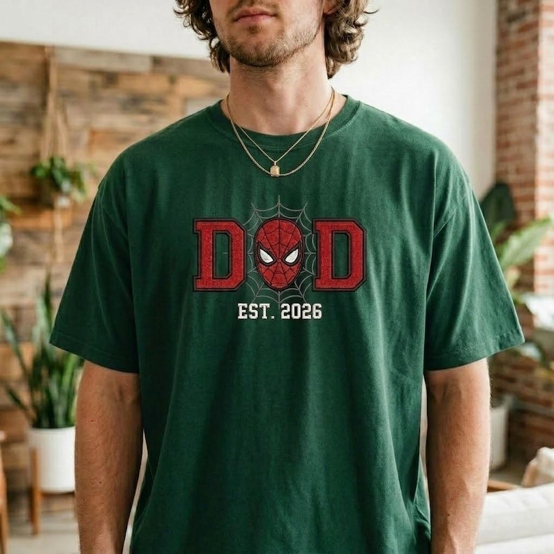 Pu&ograve; includere: T-shirt verde scuro con la scritta "DAD" in lettere rosse, con una maschera di Spider-Man al centro. Sotto il testo c'&egrave; "EST. 2026" in bianco. La maglietta &egrave; in stile casual, girocollo.