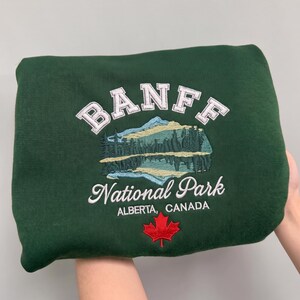May include: A dark green sweatshirt featuring an embroidered design with the text BANFF in white, followed by a mountain and forest landscape graphic, the text National Park, Alberta, CANADA, and a red maple leaf icon at the bottom.