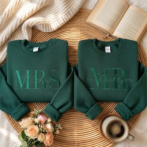 May include: Two dark green sweatshirts with embroidered text. One says "MRS Wookey" and the other says "MR Wookey". The sweatshirts are on a woven tray with a book, flowers, and a cup of coffee.