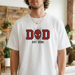 Pu&ograve; includere: T-shirt bianca con grafica rossa e nera. La grafica dice "DAD" con una maschera di Spider-Man al centro e la scritta "EST. 2026" sotto. La maglietta &egrave; realizzata in materiale morbido.