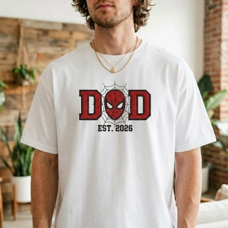 Pu&ograve; includere: T-shirt bianca con grafica rossa e nera. La grafica dice "DAD" con una maschera di Spider-Man al centro e la scritta "EST. 2026" sotto. La maglietta &egrave; realizzata in materiale morbido.