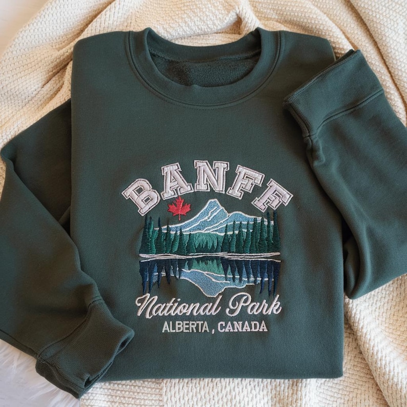 May include: A dark green crewneck sweatshirt featuring an embroidered design of Banff National Park. The graphic includes a mountain range, a forest, a lake reflection, a red maple leaf, and the text Banff National Park Alberta, Canada.