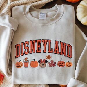 May include: Cream-colored Disneyland sweatshirt with orange lettering and fall-themed graphics. The design includes pumpkins, a Minnie Mouse pumpkin, and autumn leaves. The sweatshirt has a crew neck and long sleeves.