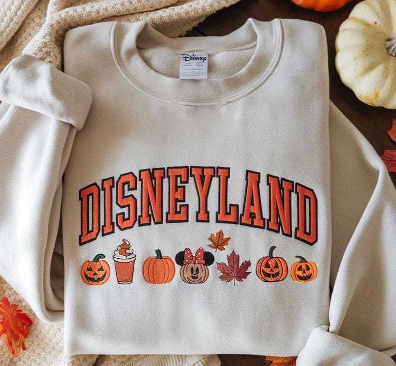 May include: Cream-colored Disneyland sweatshirt with orange lettering and fall-themed graphics. The design includes pumpkins, a Minnie Mouse pumpkin, and autumn leaves. The sweatshirt has a crew neck and long sleeves.