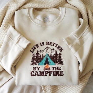May include: Cream-colored sweatshirt with the embroidered phrase "LIFE IS BETTER BY THE CAMPFIRE." The design includes a teal tent, brown trees, a campfire, and mountain silhouettes. The sweatshirt has ribbed cuffs and a crew neck.