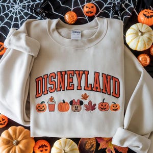 May include: Beige crewneck sweatshirt with the word "DISNEYLAND" in orange block letters. Below the text are pumpkin and leaf graphics. The sweatshirt is surrounded by pumpkins and Halloween decorations.