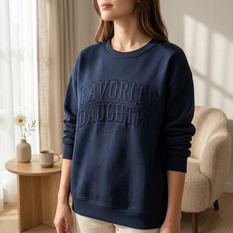 May include: Navy blue sweatshirt with the words "FAVORITE DAUGHTER" embroidered across the chest, with "SINCE 1999" underneath. The sweatshirt is a crew neck style.