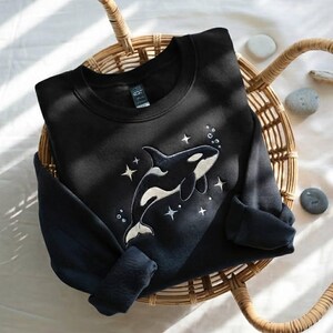 May include: A black crewneck sweatshirt featuring an embroidered design of an orca whale surrounded by white stitched stars and small bubbles. The sweatshirt has long sleeves and a ribbed collar, showcasing a detailed marine animal graphic.