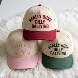 Könnte beinhalten: Drei cremefarbene Baseballkappen mit der Aufschrift "REALLY BUSY DILLY DALLYING". Die Schirme sind bordeauxrot, rosa und grün. Die Kappen sind gestapelt, um Text und Farbvarianten zu zeigen. Ein lässiges Accessoire.
