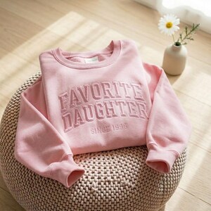 May include: A light pink sweatshirt with the embroidered text "FAVORITE DAUGHTER SINCE 1999". The crew neck sweatshirt is folded on a woven ottoman. A small vase with a daisy is in the background.