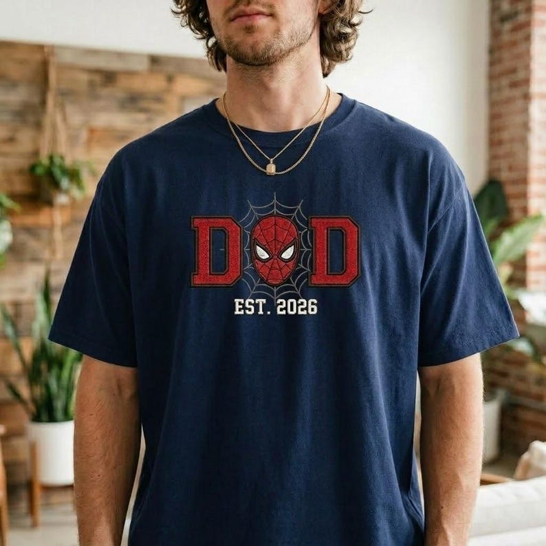 Pu&ograve; includere: T-shirt blu navy con grafica rossa e nera. La grafica presenta la parola "DAD" in lettere rosse con una maschera di Spider-Man al centro e la scritta "EST. 2026" sotto. La maglietta &egrave; a girocollo e maniche corte.