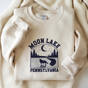 May include: Cream-colored sweatshirt with the words "MOON LAKE PENNSYLVANIA" in navy blue. The design features a crescent moon, stars, trees, and a wolf silhouette. The sweatshirt is folded on a cream-colored blanket.