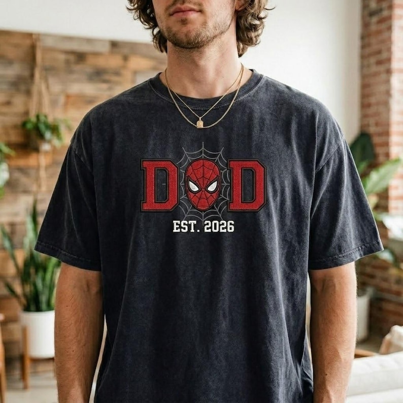 Pu&ograve; includere: T-shirt grigio scuro con grafica rossa e nera. La grafica dice "DAD" con un design a ragnatela e una maschera di Spider-Man al centro. Sotto la grafica c'&egrave; la scritta "EST. 2026".